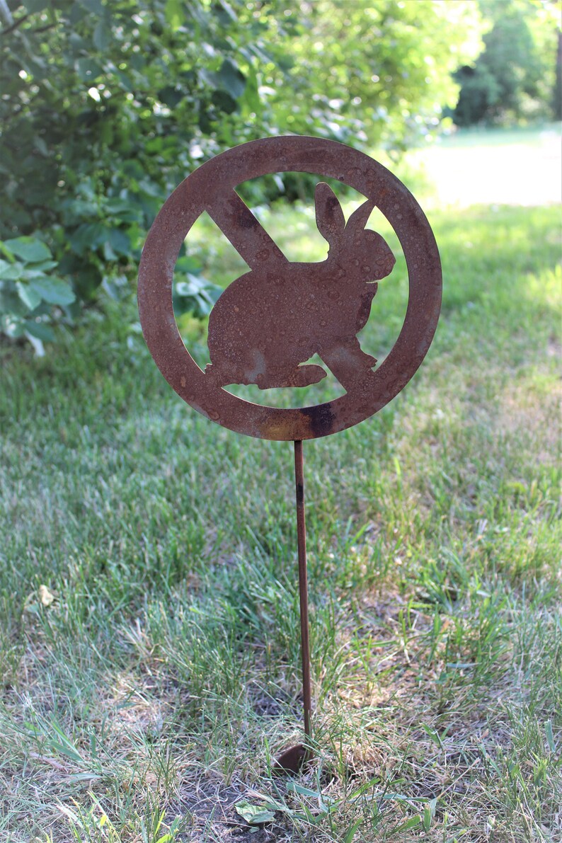 May include: A rusty metal sign with a circle and a diagonal line through it. The sign has a silhouette of a rabbit inside the circle. The sign is on a metal stake and is stuck in the ground.