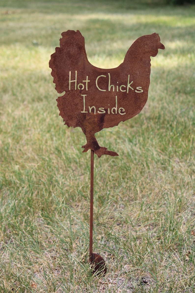 May include: A rusty metal chicken silhouette with the words "Hot Chicks Inside" written on it. The chicken is on a metal stake and is standing in a grassy field.