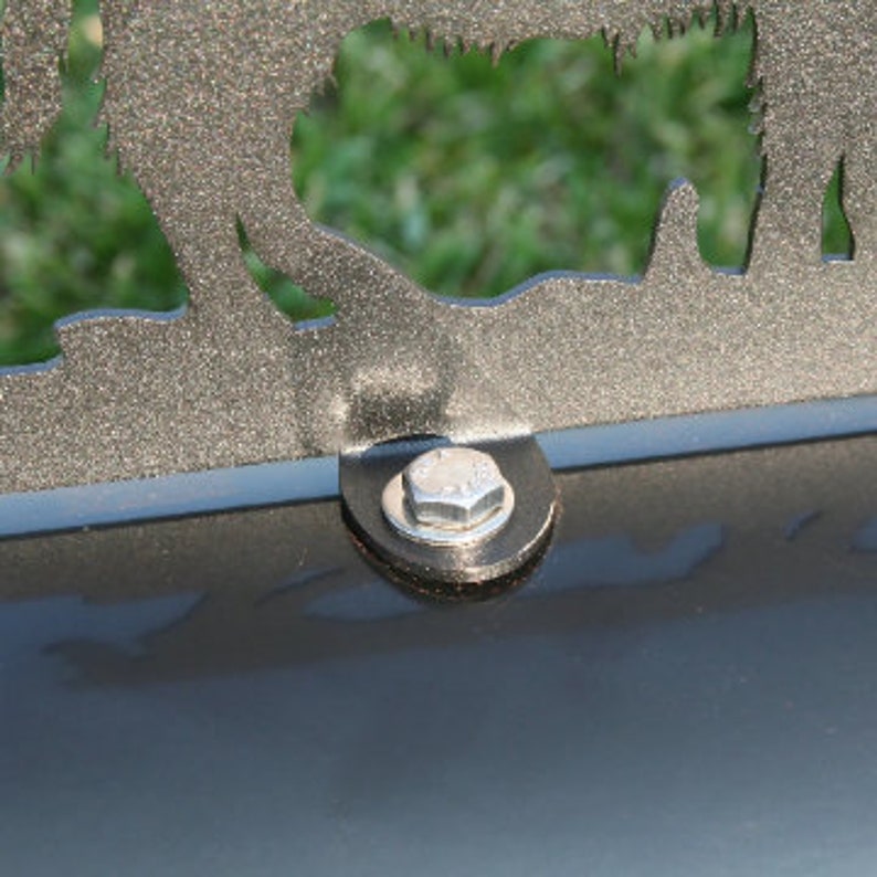 May include: Close-up of a metal decorative panel with a silhouette design of animals and mountains. The panel is attached to a black surface with a silver bolt and washer. The background is blurred green grass.