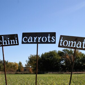 May include: Three weathered metal garden signs on stakes, each labeled with white text: "zucchini", "carrots", and "tomato". The signs are in a field with a blue sky background.