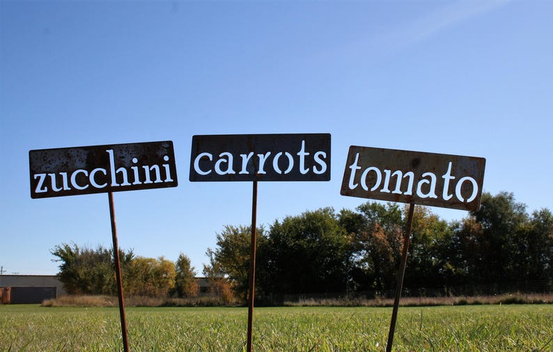 May include: Three weathered metal garden signs on stakes, each labeled with white text: "zucchini", "carrots", and "tomato". The signs are in a field with a blue sky background.