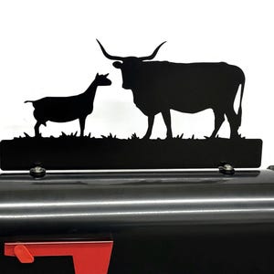 May include: Black metal mailbox with a red flag and a decorative topper featuring silhouettes of a goat and a longhorn. The mailbox is a classic design with a curved top and a front-facing door.