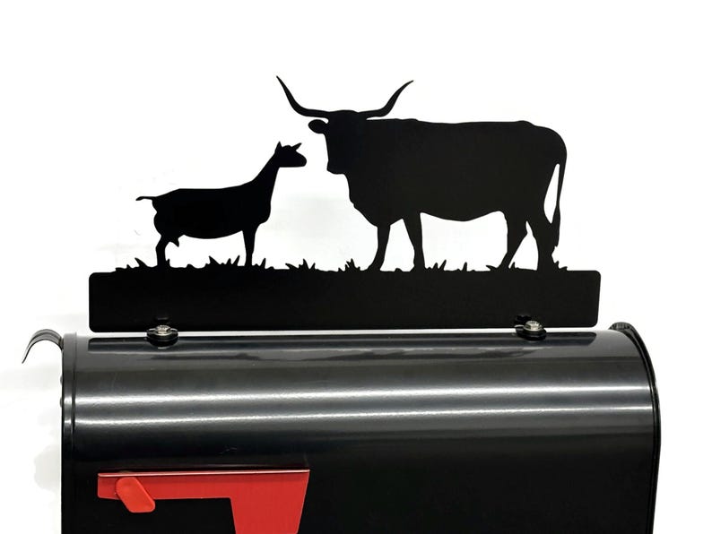 May include: Black metal mailbox with a red flag and a decorative topper featuring silhouettes of a goat and a longhorn. The mailbox is a classic design with a curved top and a front-facing door.