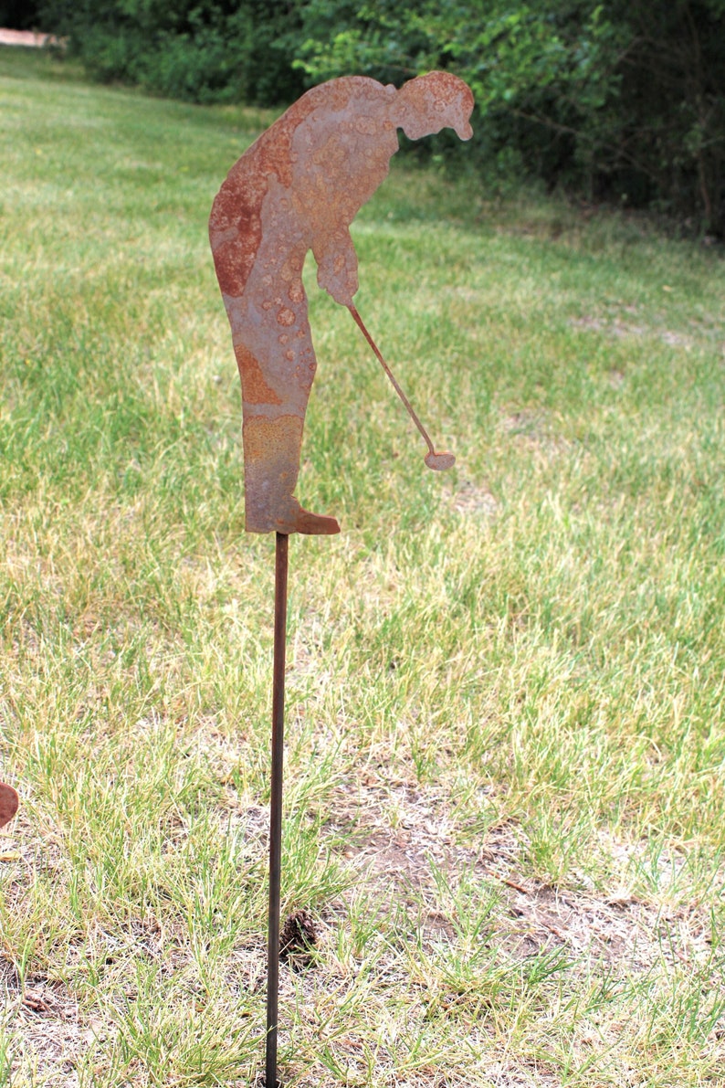 May include: A rusty metal lawn ornament depicting a golfer in a swing pose. The figure is attached to a thin metal rod for ground placement. The golfer is bent over, holding a golf club, ready to hit the ball. The ornament is set in a grassy area.