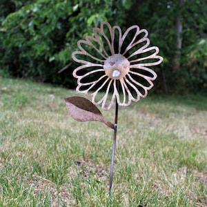 May include: A rustic metal flower garden stake with a daisy design. The flower has a weathered, brown finish and is set in a grassy yard. The flower's petals are cut out, and a small, round, clear stone is in the center.