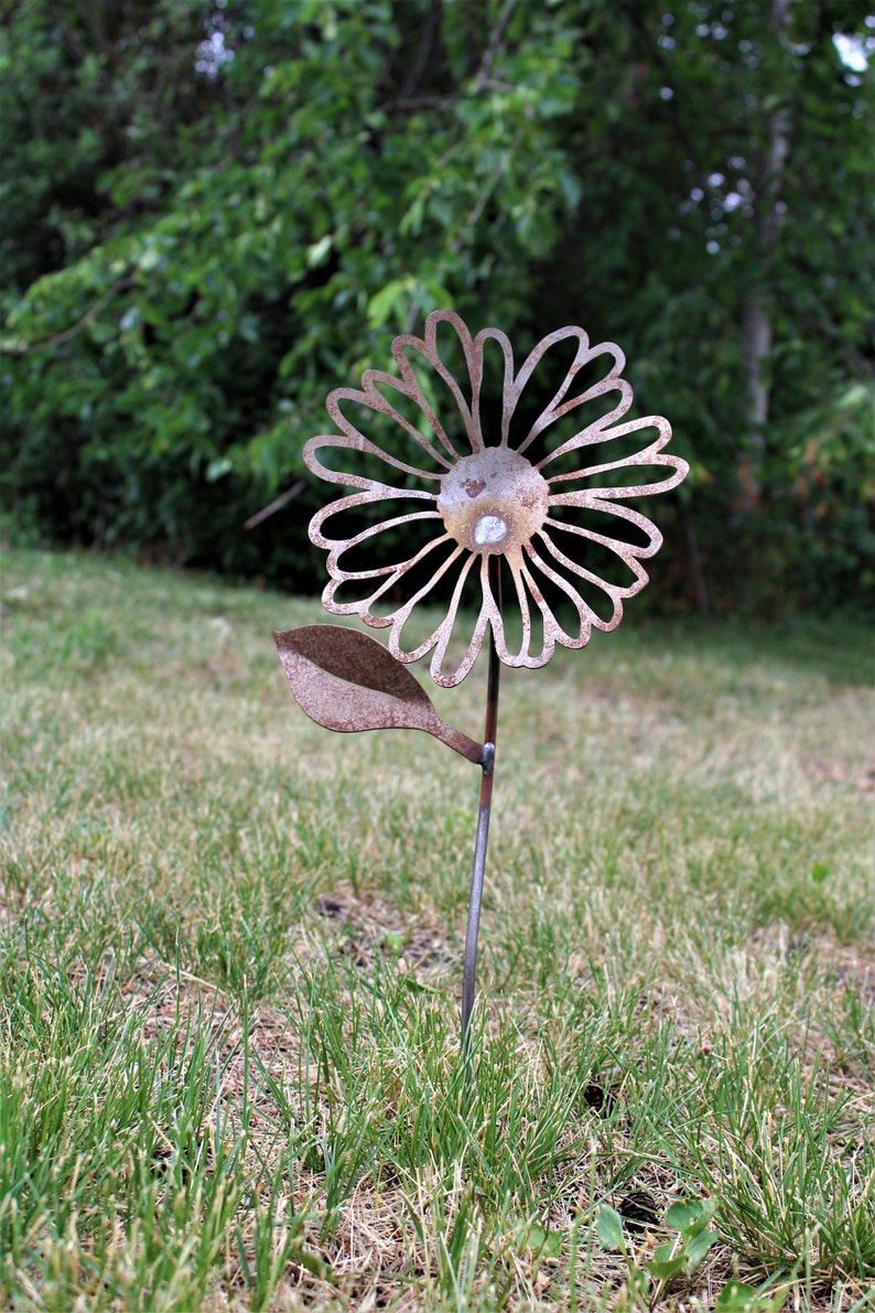 May include: A rustic metal flower garden stake with a daisy design. The flower has a weathered, brown finish and is set in a grassy yard. The flower's petals are cut out, and a small, round, clear stone is in the center.
