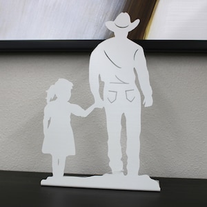May include: White metal silhouette of a father and daughter holding hands. The father is wearing a cowboy hat.