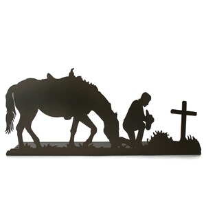 May include: A black metal silhouette depicts a horse, a kneeling figure, and a cross. The horse is saddled and grazing. The figure is kneeling with a hat. The cross stands on a grassy base.