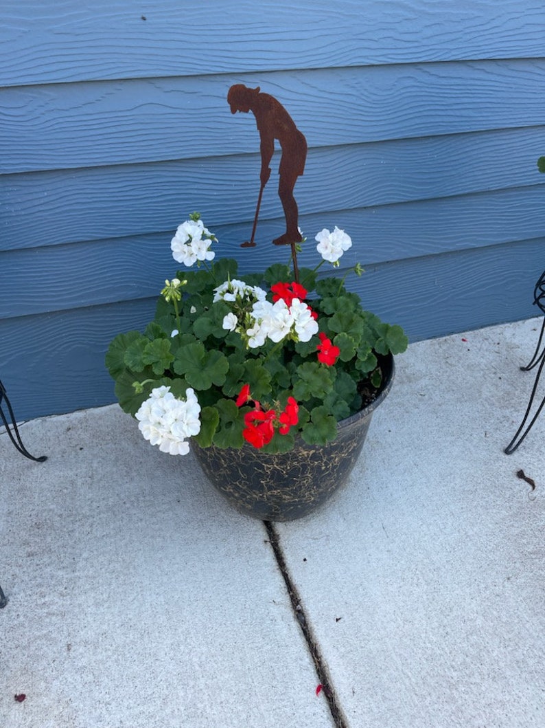 May include: A black and gold decorative planter filled with white and red geraniums. A rusty metal silhouette of a golfer is placed in the center of the planter. The planter is sitting on a concrete surface.