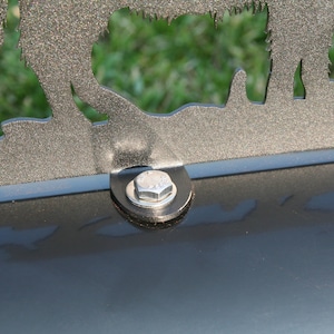 May include: A black metal decorative piece featuring a wolf silhouette, secured to a black cylindrical object with a bolt and washer. The background shows blurred green grass.