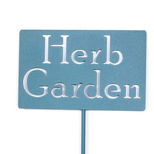 May include: A teal metal garden stake with the words "Herb Garden" cut out in white. The rectangular sign is mounted on a thin, matching teal pole, perfect for marking a herb garden.