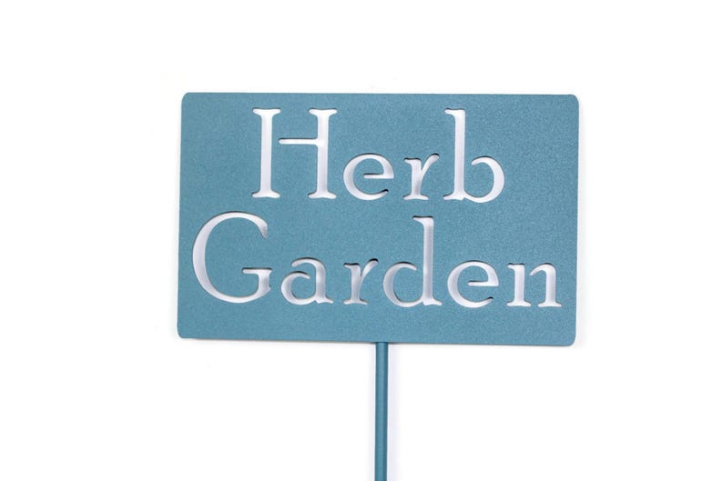 May include: A teal metal garden stake with the words "Herb Garden" cut out in white. The rectangular sign is mounted on a thin, matching teal pole, perfect for marking a herb garden.