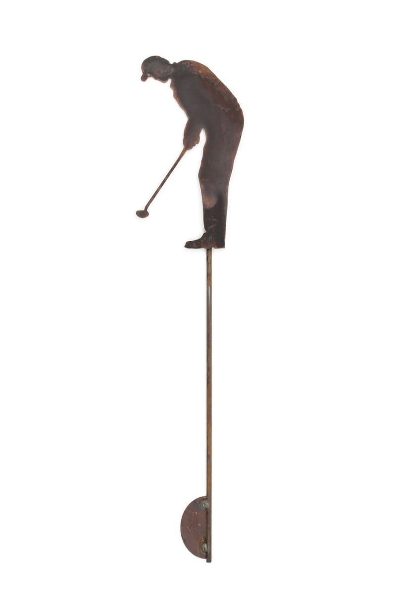 May include: A silhouette of a golfer in a rusty metal finish, poised to swing. The figure is attached to a long, thin rod, with a circular base. This outdoor decor piece is designed for garden or yard display.