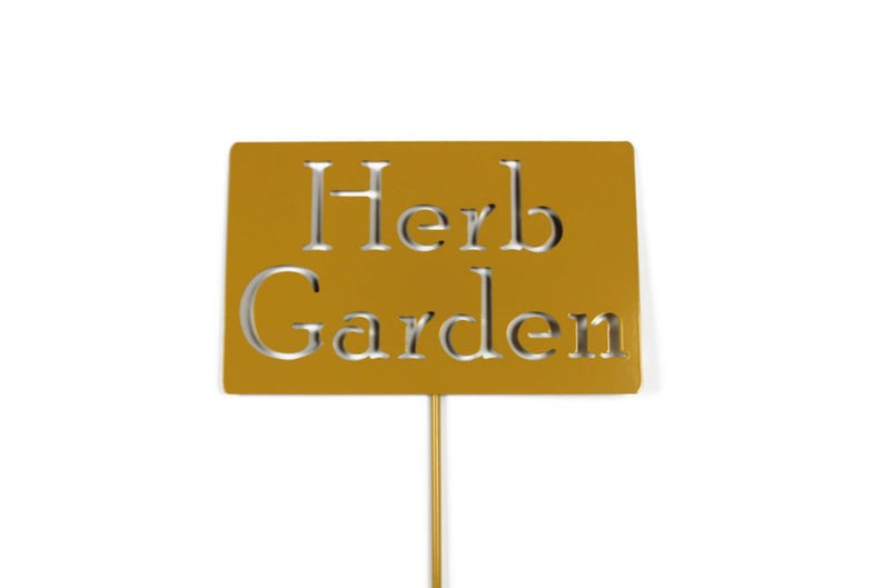 May include: A golden-yellow metal garden stake with the words "Herb Garden" cut out. The sign is rectangular and mounted on a thin, gold-colored rod. Ideal for marking herbs in a garden.