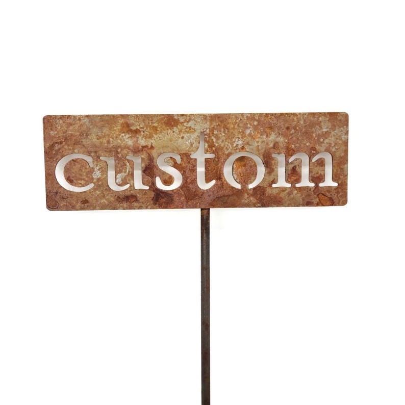 May include: A rectangular, weathered metal garden stake with the word "custom" cut out. The stake has a rusty brown and orange patina, and is mounted on a dark metal rod. The background is white.