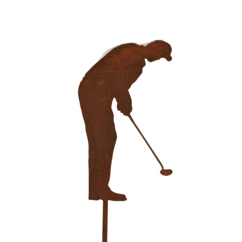 May include: A rusty brown metal silhouette of a golfer in a putting stance. The figure is bent over, holding a golf club, with a cap on their head. The silhouette is mounted on a stake for garden or yard decor.