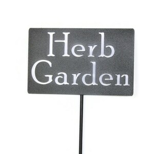 May include: A rectangular, dark gray garden sign with the words "Herb Garden" in white, sans-serif font. The sign is mounted on a thin, black metal stake, suitable for outdoor use in a garden or yard.