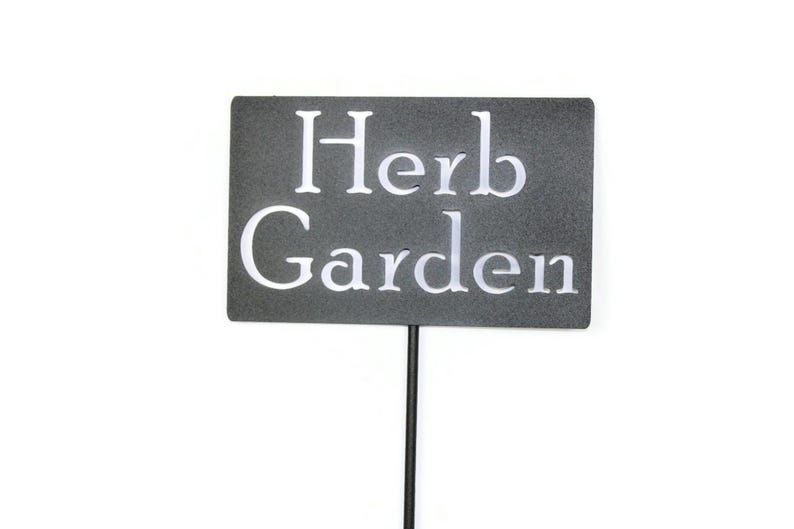 May include: A rectangular, dark gray garden sign with the words "Herb Garden" in white, sans-serif font. The sign is mounted on a thin, black metal stake, suitable for outdoor use in a garden or yard.
