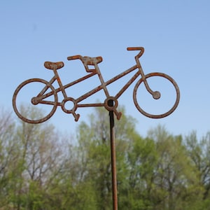 May include: A rusty metal silhouette of a tandem bicycle, mounted on a metal pole. The bicycle is against a blue sky with trees in the background.