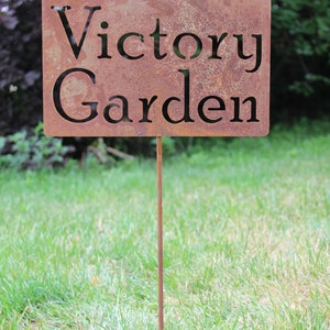 May include: A rectangular, weathered metal garden sign with the words "Victory Garden" cut out. The sign is mounted on a thin, rusty metal stake and is placed in a grassy area.