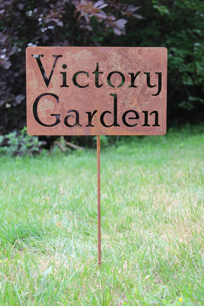 May include: A rectangular, weathered metal garden sign with the words "Victory Garden" cut out. The sign is mounted on a thin, rusty metal stake and is placed in a grassy area.