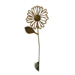 May include: A metal daisy garden stake with a rusty brown finish. The flower has a detailed openwork design, a central disc, and a single leaf. The stem is slender and extends to a rounded base for ground placement.