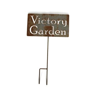 May include: A rectangular, weathered metal garden stake with the words "Victory Garden" cut out. The sign is mounted on a thin, dark metal rod with a two-pronged base for easy placement in the ground. The sign has a rustic, aged appearance.