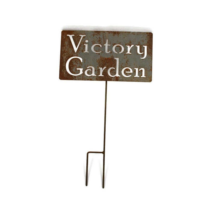 May include: A rectangular, weathered metal garden stake with the words "Victory Garden" cut out. The sign is mounted on a thin, dark metal rod with a two-pronged base for easy placement in the ground. The sign has a rustic, aged appearance.