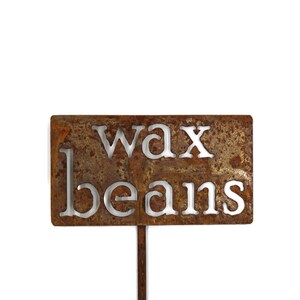 May include: A rectangular, rusty metal garden stake with the words "wax beans" cut out. The stake is designed for labeling plants in a garden or vegetable patch. The metal has a weathered, aged appearance.