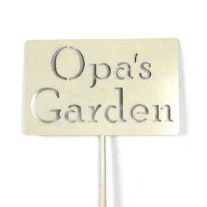 May include: A cream-colored garden sign with the words "Opa's Garden" cut out. The sign is rectangular and mounted on a thin, white metal stake. The text is in a simple, sans-serif font, suitable for outdoor use.