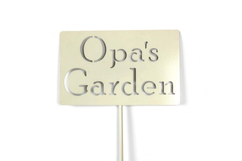 May include: A cream-colored garden sign with the words "Opa's Garden" cut out. The sign is rectangular and mounted on a thin, white metal stake. The text is in a simple, sans-serif font, suitable for outdoor use.