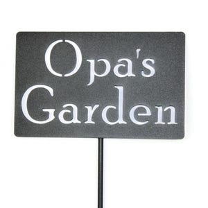May include: A rectangular garden sign with the words "Opa's Garden" in white, sans-serif font. The sign is dark gray and mounted on a thin, black metal stake, suitable for outdoor use.