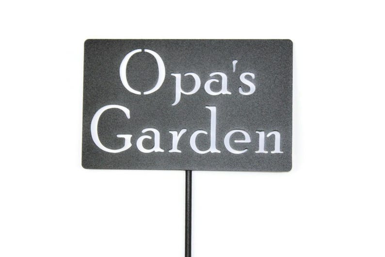 May include: A rectangular garden sign with the words "Opa's Garden" in white, sans-serif font. The sign is dark gray and mounted on a thin, black metal stake, suitable for outdoor use.