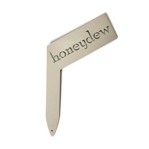 May include: A metal garden marker with the word "honeydew" cut out. The marker is a light beige color and has a pointed end.