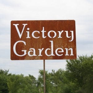 May include: A rectangular, weathered metal sign with the words "Victory Garden" cut out in white lettering. The sign is mounted on a thin metal pole, set against a backdrop of green trees and a cloudy sky.