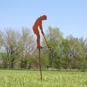 May include: A rusty metal lawn ornament depicting a golfer in mid-swing. The silhouette is mounted on a long stake, positioned in a grassy field with trees and a blue sky in the background. A decorative garden accent.