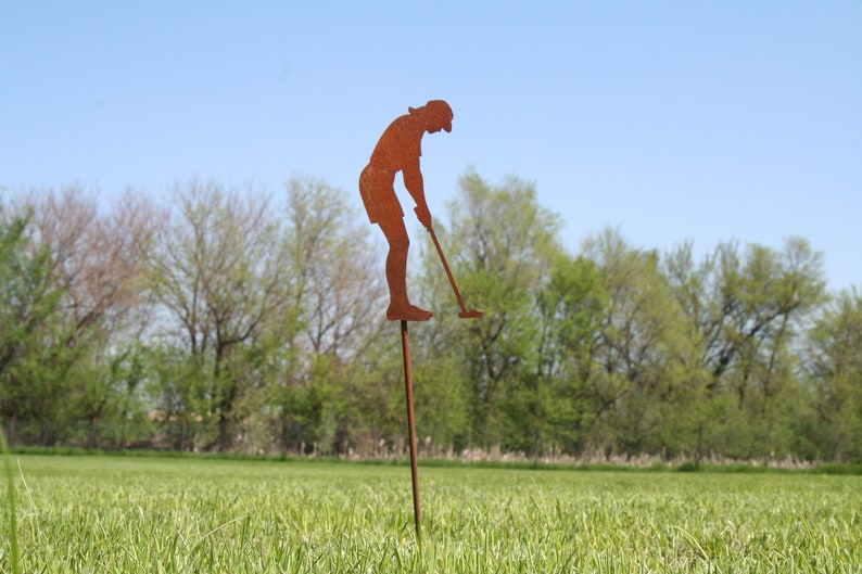 May include: A rusty metal lawn ornament depicting a golfer in mid-swing. The silhouette is mounted on a long stake, positioned in a grassy field with trees and a blue sky in the background. A decorative garden accent.