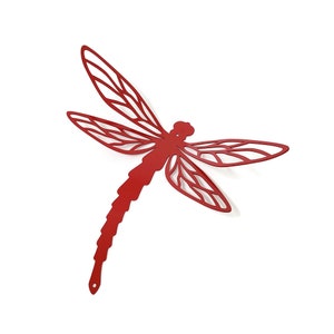 May include: A red metal dragonfly wall art piece. The dragonfly has intricate cut-out wing designs and a long, segmented body. The wings are spread wide, and the piece has a small hole at the end of the tail for hanging. The dragonfly is a vibrant red color.