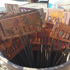 May include: A collection of weathered metal garden markers with cut-out lettering. The markers, displaying names like "beans," "tomato," and "garlic," are arranged in a terracotta pot. The metal has a rustic, orange-brown patina.