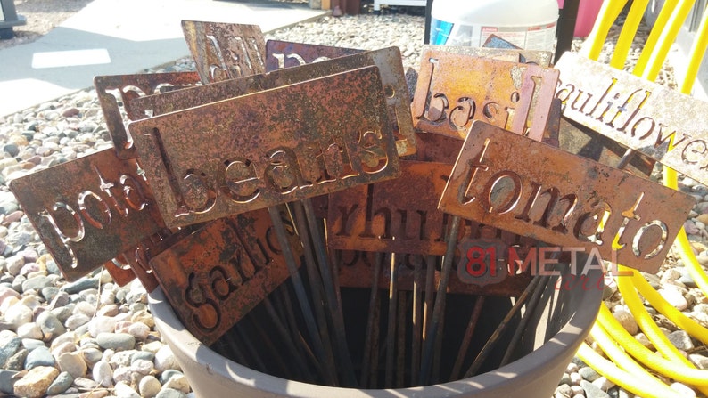 May include: A collection of weathered metal garden markers with cut-out lettering. The markers, displaying names like "beans," "tomato," and "garlic," are arranged in a terracotta pot. The metal has a rustic, orange-brown patina.
