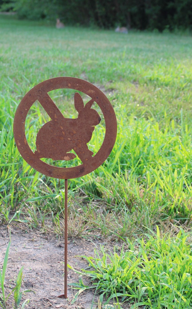 May include: A rusty metal sign with a circle and a diagonal line through it. Inside the circle is a silhouette of a rabbit. The sign is on a metal stake and is stuck in the ground.
