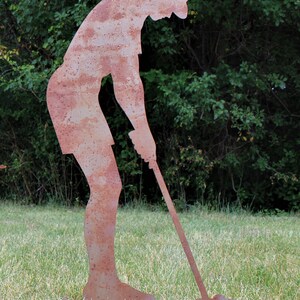 May include: A rusty metal silhouette of a golfer in a putting stance. The figure is on a grassy lawn with a backdrop of green foliage. The golfer is bent over, holding a golf club, ready to swing.