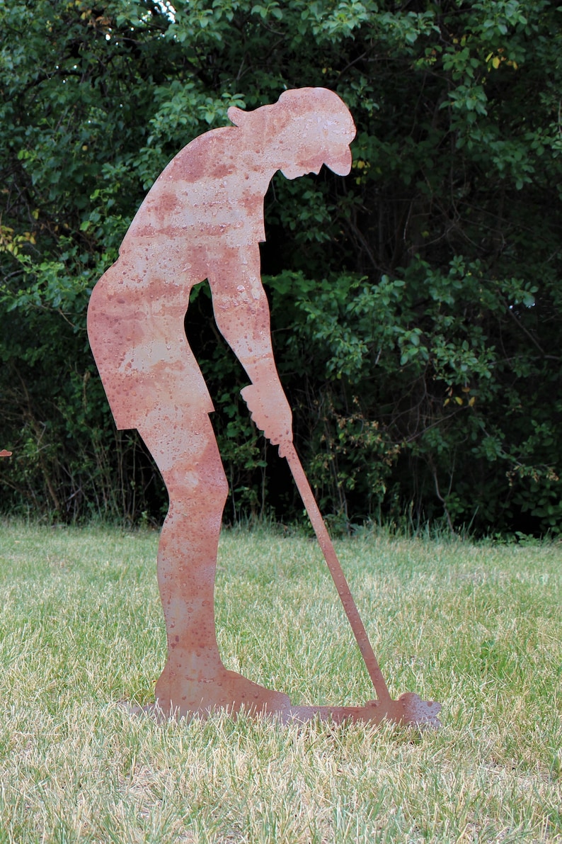 May include: A rusty metal silhouette of a golfer in a putting stance. The figure is on a grassy lawn with a backdrop of green foliage. The golfer is bent over, holding a golf club, ready to swing.