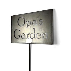 May include: A rectangular metal garden sign with the words "Opa's Garden" cut out. The sign is mounted on a black metal stake. The sign has a weathered, metallic finish, and is set against a white background.
