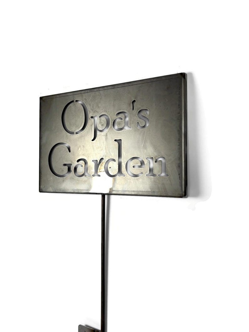 May include: A rectangular metal garden sign with the words "Opa's Garden" cut out. The sign is mounted on a black metal stake. The sign has a weathered, metallic finish, and is set against a white background.
