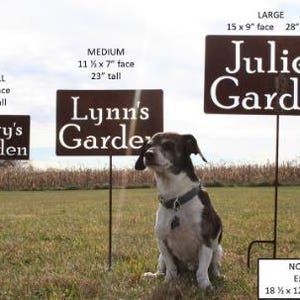May include: Three metal garden signs with the names "Mary's Garden", "Lynn's Garden", and "Julie's Garden" are displayed in a grassy field. The signs vary in size, with the largest measuring 15 x 9 inches. A dog sits in front of the signs.