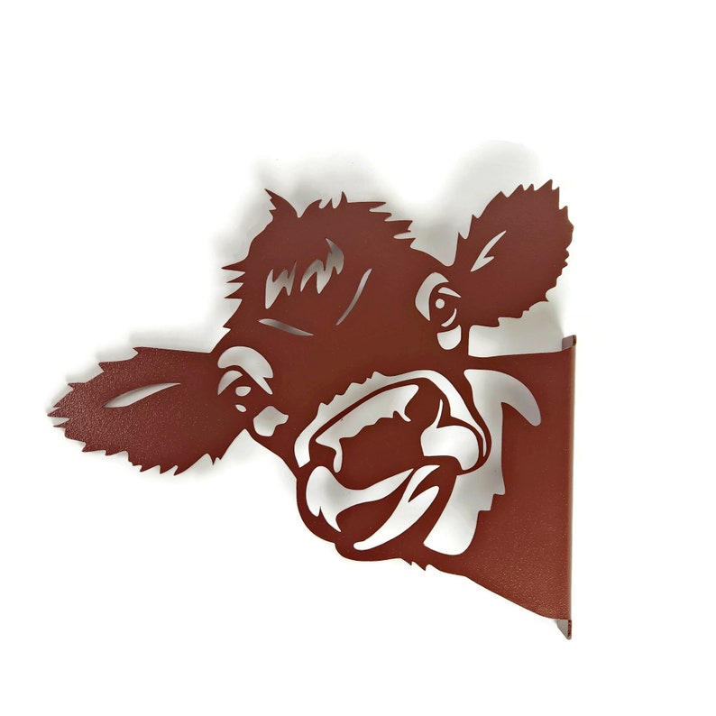 May include: A brown metal cutout of a cow's head with an open mouth and a tongue sticking out. The cow's head is facing the right side of the image and is partially obscured by a brown metal background.