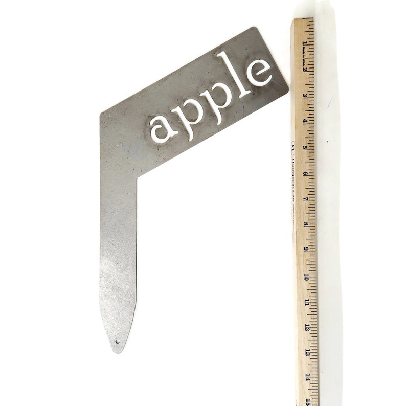 May include: A metal garden marker with the word "apple" cut out in white lettering. The marker is shaped like a right angle with a pointed end.