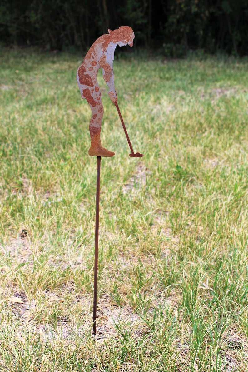 May include: A rusty metal golfer silhouette, with a golf club, is staked into the ground. The figure is in a putting stance, with a weathered, textured appearance. The sculpture is set in a grassy field.