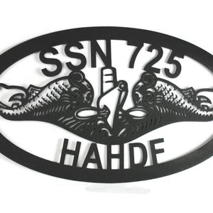 Puede incluir: Un letrero de pared ovalado de metal negro con un dise&ntilde;o recortado de la insignia de guerra submarina de la Marina de los EE. UU., tambi&eacute;n conocida como delfines. El texto SSN 725 est&aacute; arriba y el texto HAHDF est&aacute; abajo del &oacute;valo.